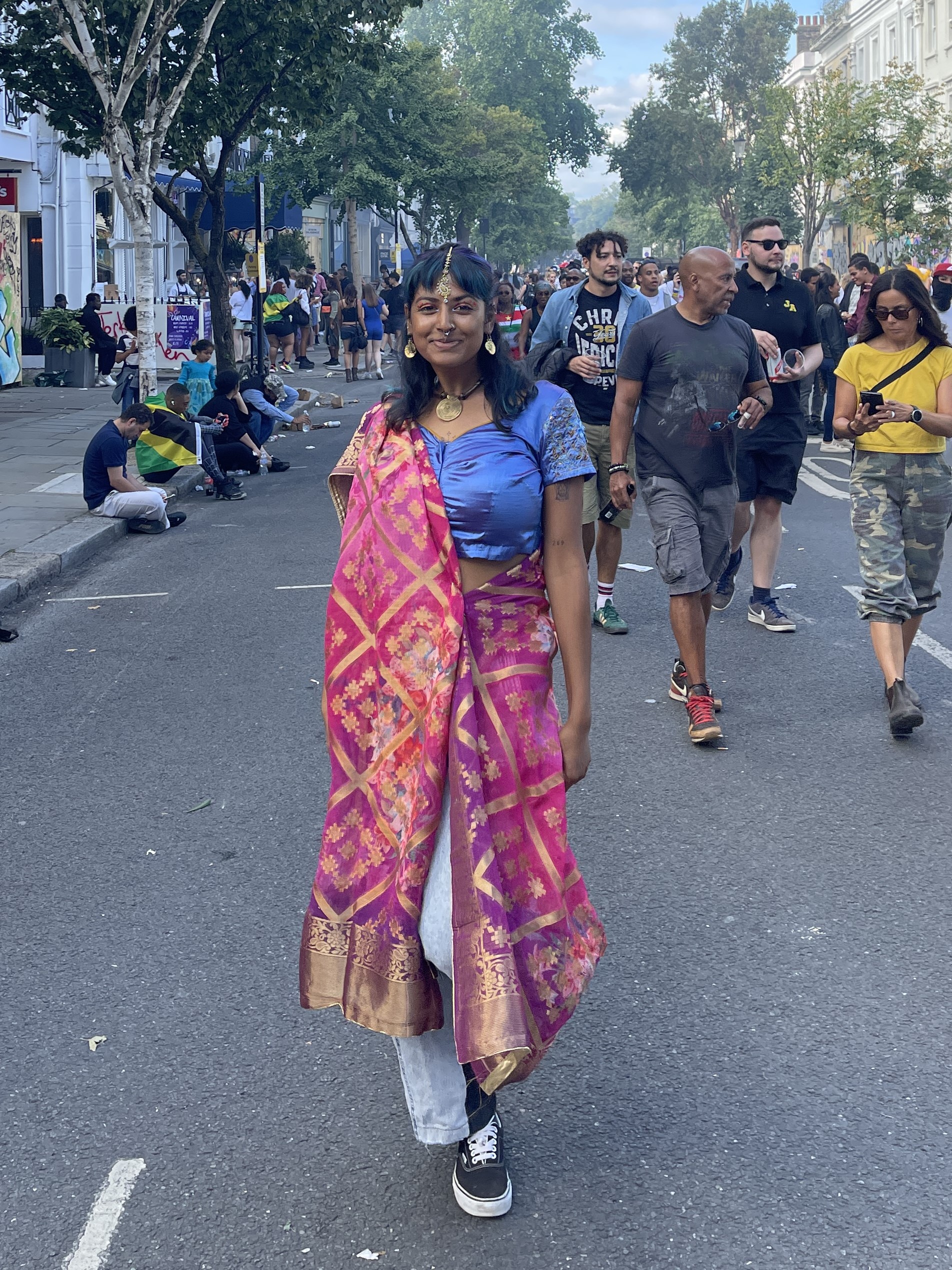 Foto von Kantom Azad auf einer Straße, umgeben von Menschen. Sie trägt Sneaker, Jeans und einen bunten Sari sowie traditionellen Schmuck.