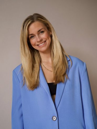 Lara Sophie Bothur in einem hellblauten Blazer im Porträt, blonde lange Haare, sie legt den Kopf leicht schief und lächelt in die Kamera.