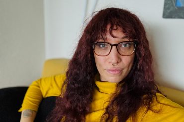 Das Foto zeigt Jasmin Dickerson. Sie hat lange rotbraune Haare, trägt eine Brille, einen gelben Pullover und sitzt auf einer gelben Couch.