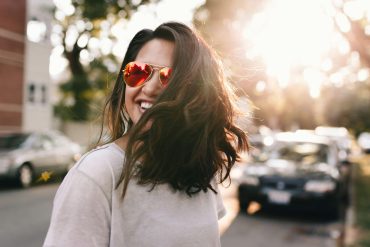 Frau mit roter Sonnenbrille und langem dunklem Haar lacht auf der Straße, sie wirkt glücklich.