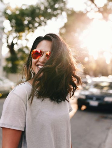 Frau mit roter Sonnenbrille und langem dunklem Haar lacht auf der Straße, sie wirkt glücklich.