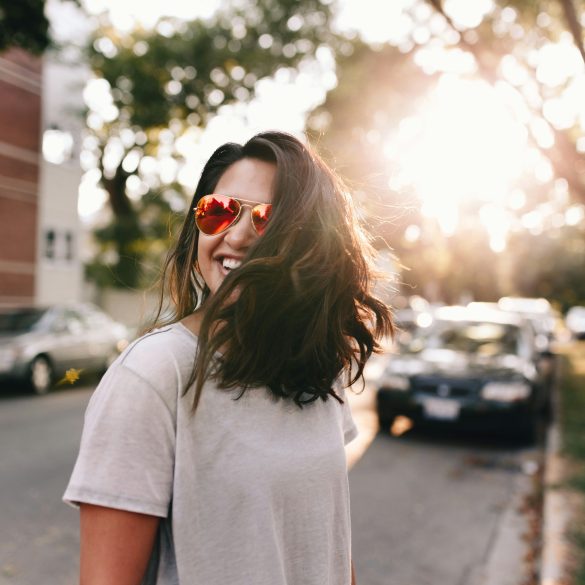 Frau mit roter Sonnenbrille und langem dunklem Haar lacht auf der Straße, sie wirkt glücklich.