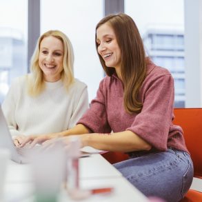 Zwei Frauen am Schreibtisch. Beide lachen, eine hat blonde, eine brünettfarbene Haarre. Im Hintergrund ein Regal und ein Fenster oberhalb einer größeren Stadt.