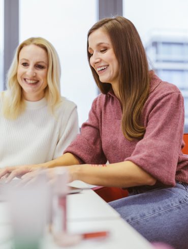 Zwei Frauen am Schreibtisch. Beide lachen, eine hat blonde, eine brünettfarbene Haarre. Im Hintergrund ein Regal und ein Fenster oberhalb einer größeren Stadt.