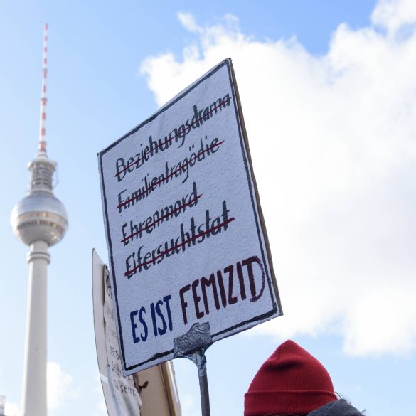 Demonstrationsschild: Es ist Femizid. Hintergrund: Blauter Himmel und Fernsehturm in Berlin