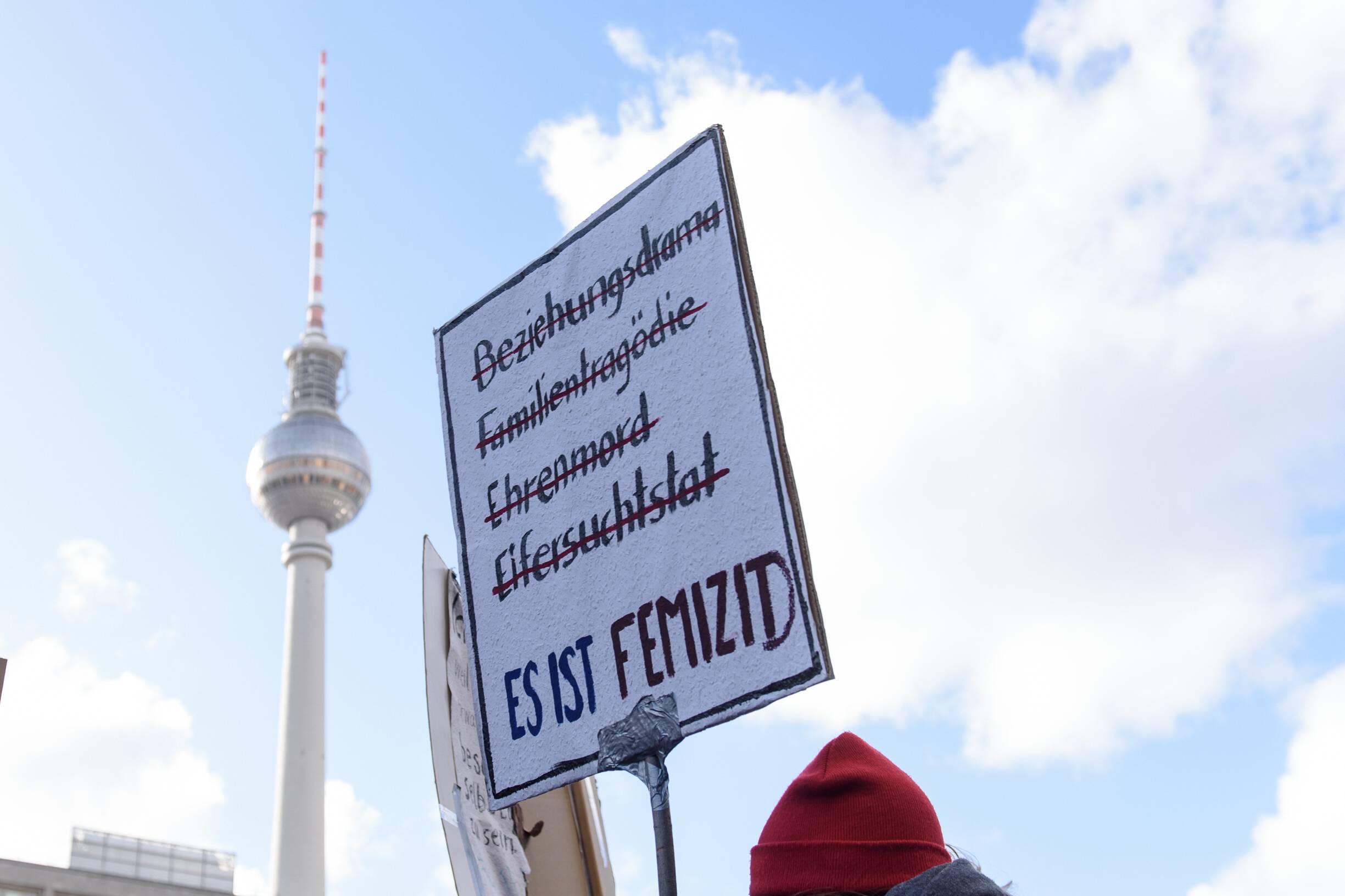 Demonstrationsschild: Es ist Femizid. Hintergrund: Blauter Himmel und Fernsehturm in Berlin