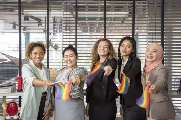 Fünf Menschen stehen nebeneinander, lächeln und halten Regenbogenfahnen in den Händen