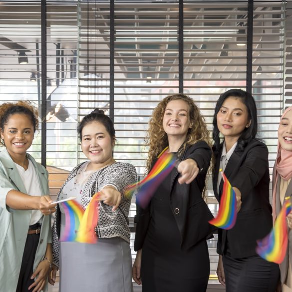 Fünf Menschen stehen nebeneinander, lächeln und halten Regenbogenfahnen in den Händen