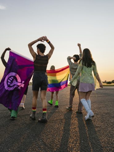 Vier Menschen mit dem Rücken zur Kamera gewandt vor einem Sonnenuntergang. Eine Person hält eine Regenbogenflagge und eine andere hält eine Feminismus-Flagge,