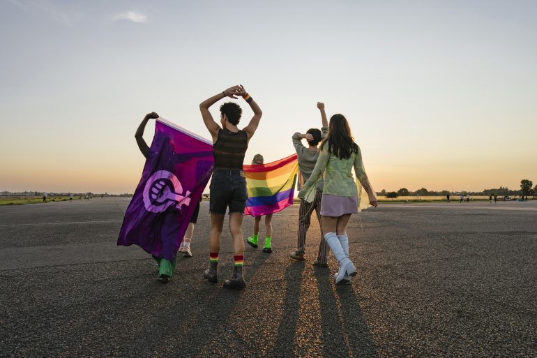 Vier Menschen mit dem Rücken zur Kamera gewandt vor einem Sonnenuntergang. Eine Person hält eine Regenbogenflagge und eine andere hält eine Feminismus-Flagge,