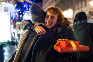 Das Foto zeigt zwei Frauen, die sich umarmen. Im Hintergrund ist ein Gebäude und eine Lichterkette zu sehen. Die Frau rechts hält in rot verpackte Geschenke und lächelt.