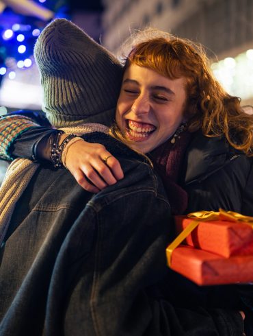 Das Foto zeigt zwei Frauen, die sich umarmen. Im Hintergrund ist ein Gebäude und eine Lichterkette zu sehen. Die Frau rechts hält in rot verpackte Geschenke und lächelt.