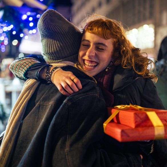 Das Foto zeigt zwei Frauen, die sich umarmen. Im Hintergrund ist ein Gebäude und eine Lichterkette zu sehen. Die Frau rechts hält in rot verpackte Geschenke und lächelt.