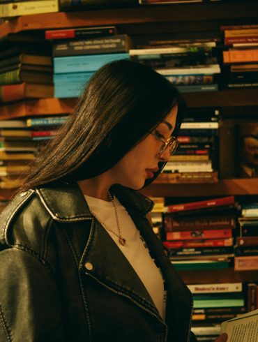 Junge Frau steht vor einem riesigen Bücherregal und liest eines der bunten Bücher. Sie trägt eine Brille und langes schwarzes Haar.