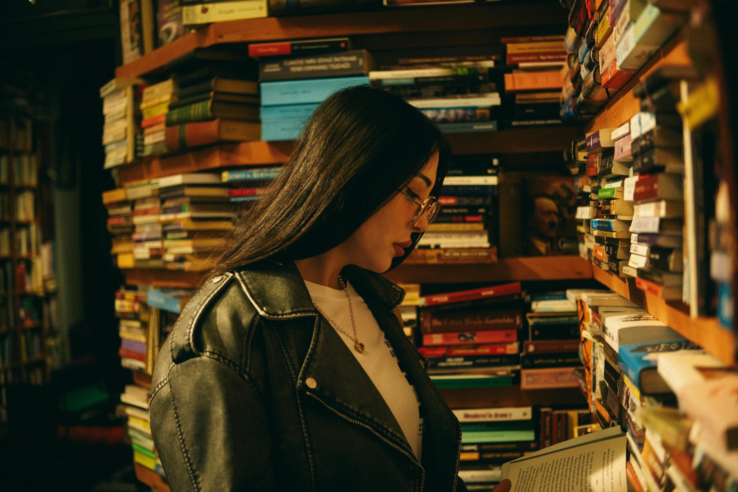 Junge Frau steht vor einem riesigen Bücherregal und liest eines der bunten Bücher. Sie trägt eine Brille und langes schwarzes Haar.