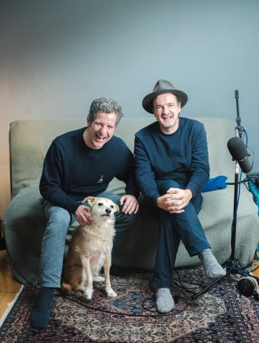 Die Podcast-Hosts Winson und Ueli sitzen im Aufnahme-Wohnzimmer, vor ihnen ein goldbrauner Hund, mittelgroß. Sie lachen in die Kamera.