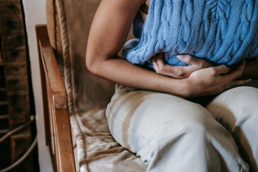 Frau hält sich den Bauch, trägt blaues armfreies gestricktes Shirt und eine beige Hose, Kopf ist nicht zu erkennen.