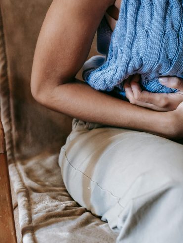 Frau hält sich den Bauch, trägt blaues armfreies gestricktes Shirt und eine beige Hose, Kopf ist nicht zu erkennen.