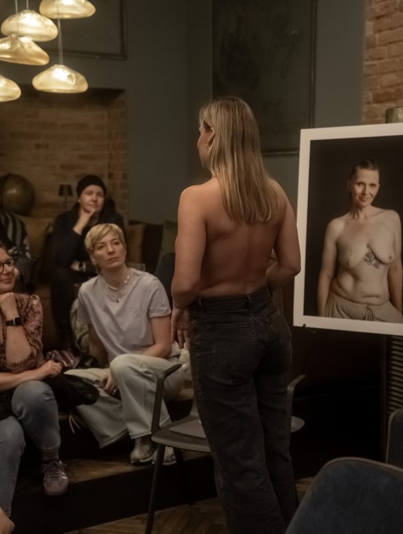 Eine Frau zeigt ihre Brustkrebsnarbe. Rundherum stehen Bilder von Frauen, die ihre Narben fotografieren ließen.