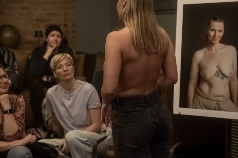 Eine Frau zeigt ihre Brustkrebsnarbe. Rundherum stehen Bilder von Frauen, die ihre Narben fotografieren ließen.