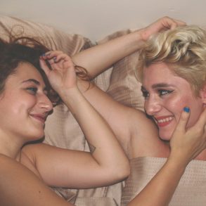 Zwei Frauen liegen nebeneinander im Bett, lächeln sich an. Sie sind nackt und berühren sich sanft mit den Armen.