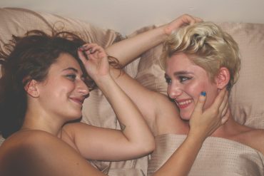 Zwei Frauen liegen nebeneinander im Bett, lächeln sich an. Sie sind nackt und berühren sich sanft mit den Armen.