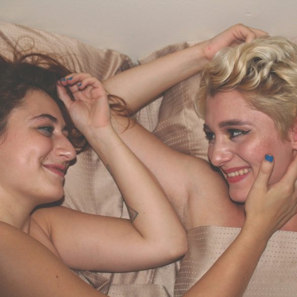 Zwei Frauen liegen nebeneinander im Bett, lächeln sich an. Sie sind nackt und berühren sich sanft mit den Armen.