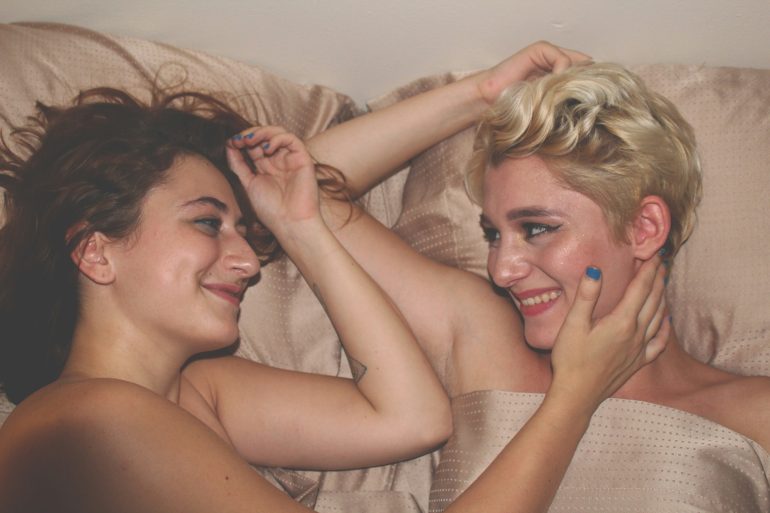 Zwei Frauen liegen nebeneinander im Bett, lächeln sich an. Sie sind nackt und berühren sich sanft mit den Armen.
